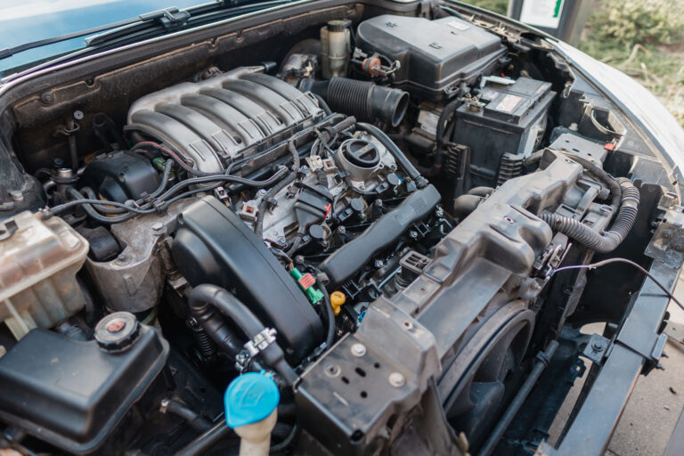 Open car engine bay showing visible V6 engine, intake manifold, hoses, wiring, battery, coolant reservoir, radiator support and detailed mechanical components in natural daylight